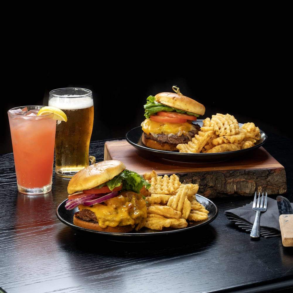 A couple of plates with burgers, waffle fries,  a cocktail and a glass of beer on a table